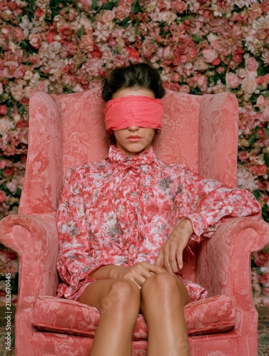 A young woman sits in an English armchair against a backdrop of flowers, her eyes hidden by a blindfold 