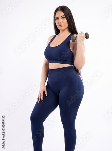 Confident curvy woman in navy blue activewear holding a dumbbell on a white studio background