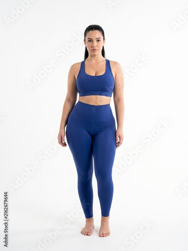 Confident curvy woman standing in a full-length pose wearing a matching blue activewear set against a clean white studio background