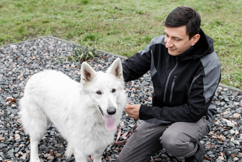 Smiling man crouching next to his White Swiss Shepherd dog on a pebble ground outdoor. Male owner petting and holding his white fluffy pet on a leash during a walk. Sport concept.