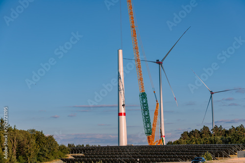 Construction of wind turbine with heavy mobile crane and solar panel farm underneath against clear blue sky, renewable hybrid energy concept