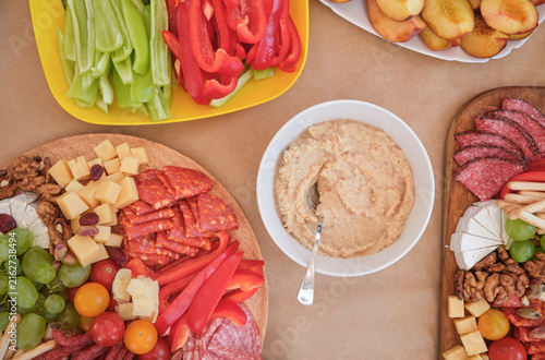 Top view of creamy hummus surrounded by colorful vegetables, cheese, grapes and assorted savory snacks. Abundant party food arrangement for gatherings and entertaining