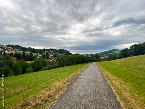 Asphalt path through green meadow leading towards rural village and forested hills