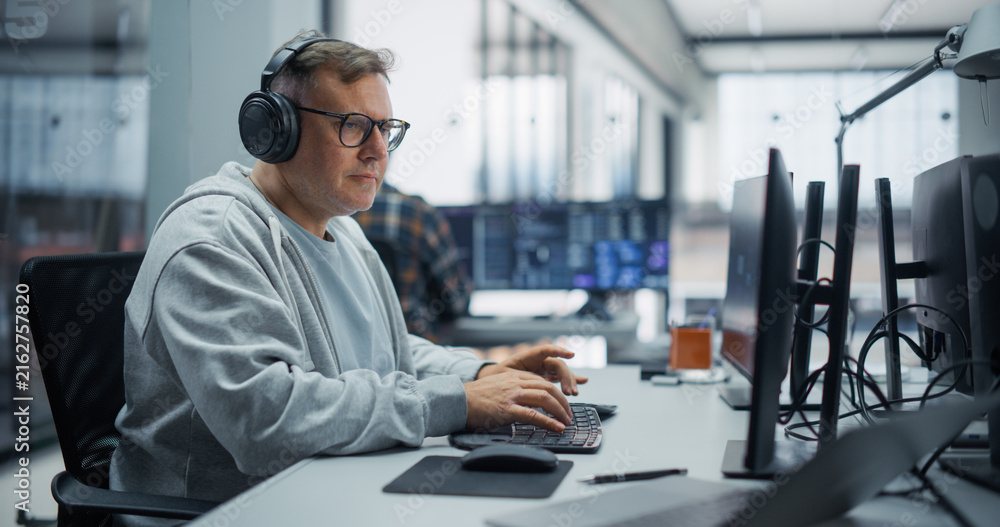 Fototapeta premium Portrait of a Concentrated Middle Aged Caucasian Male Software Developer in Glasses and Headphones Writing Program Code on a Desktop Computer, Building Applications With a Diverse Team in an Office.