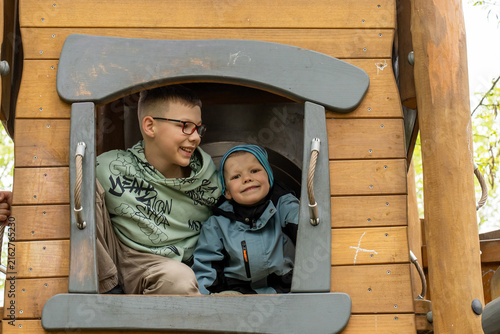Happy brothers sitting close together inside wooden playground tower, older boy looking affectionately at younger sibling. Concept of brotherly love, emotional connection, trust and family bonding. 