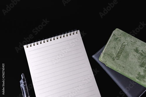 Blank lined spiral notepad pen and stacked closed notebooks on black background