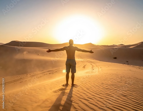 Man Standing on Desert Dune Embracing Sunset with Open Arms and Bright Horizon in Background
