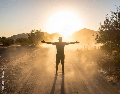Man Embraces Sunset in Desert Landscape with Dusty Trail and Mountain Silhouette