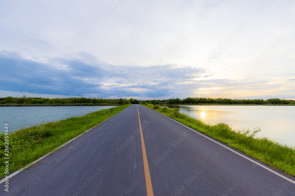 Fototapeta premium Asphalt Road Through Coastal Mangrove Forest at Sunset,An empty, scenic asphalt road stretches straight through a coastal aquaculture area and mangrove forest towards the horizon, captured during the