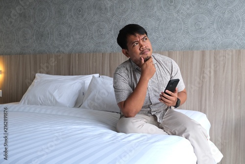 Canvastavla Thoughtful Asian man sitting on a bed, holding a smartphone and touching his chin while looking upward and considering an idea