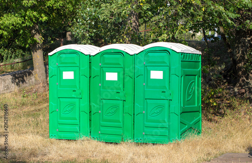 Photography Three green portable toilets with white roofs stand together on dry grass in a s