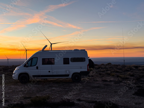 A white camper van parked on a remote ridge silhouette against a dramatic orange and purple sunset sky with distant wind turbines.