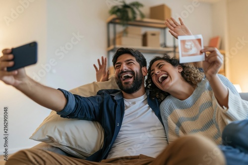 Excited couple sitting on a sofa holding a phone and ultrasound scan, laughing naturally during a video call for pregnancy announcement and family marketing
