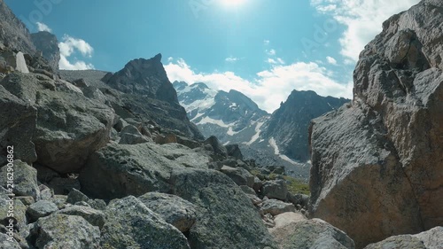 Timelapse of fast moving clouds over mountains. Sunlit alpine scene framed by rugged boulders leading to snow capped peaks under clear blue sky