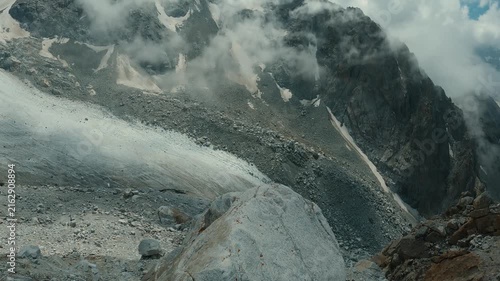 Glacier flows down rocky valley surrounded by snow capped peaks under partly cloudy sky. Cool tones dominate with white ice, gray rock, blue sky. Majestic alpine landscape evokes adventure solitude