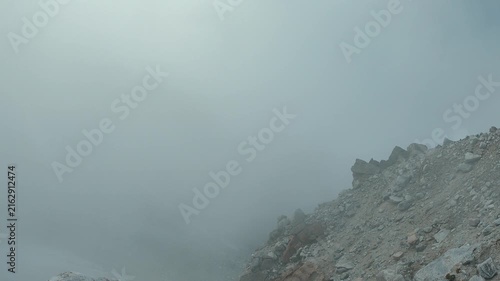 Dramatic mountain landscape with jagged peaks partially obscured by low clouds. Rocky slope in foreground under bright blue sky. Cool tones, natural lighting, moody atmosphere