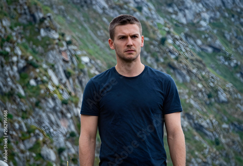 Outdoor adventure and exploration, serious young man in a dark t-shirt standing against a rugged mountain landscape