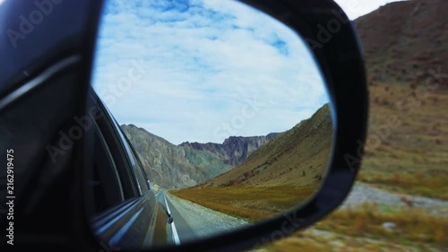 View through car side mirror shows winding mountain road. Mountains stretch under blue sky with clouds. Road curves through rocky terrain with grassy slopes. Scene evokes freedom, adventure