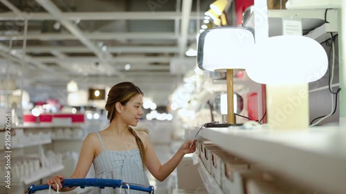 Happy young Asian woman pushing a shopping cart while carefully choosing table lamps in a large retail furniture store. She checks the price tag to buy new lighting for her modern home interior.