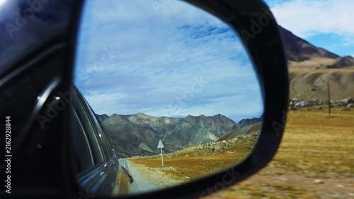 Car side mirror reflects mountain road under blue sky. Journey through scenic highlands with winding road. Serene view evokes freedom and adventure. Ideal for travel, road trip