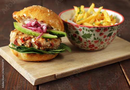 Gourmet chicken burger with avocado, pickled red onion, spinach, and crispy french fries on wooden background