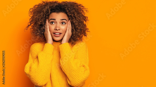 Surprised young woman with curly hair holding hands on face on orange background