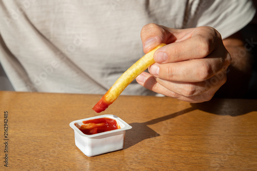 a portioned sauce boat (dip box) with tomato ketchup. Man dipping a potato fry into ketchup sauce at a wooden table. Casual fast food snack and savory eating 