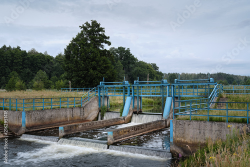 Wide view of a multi-gate weir with blue sluice gates and water flowing through concrete channels, illustrating river engineering and water management.