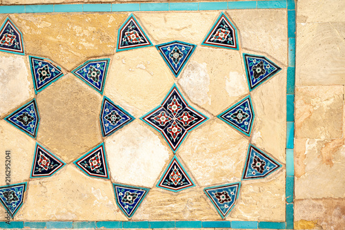 Ceramic tile pattern on a stone wall with blue triangular ornaments, floral motifs and turquoise borders in a geometric arrangement