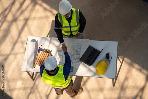 Top view of two Asian construction engineers shaking hands over a table with blueprints, a laptop, and material samples at a building site.