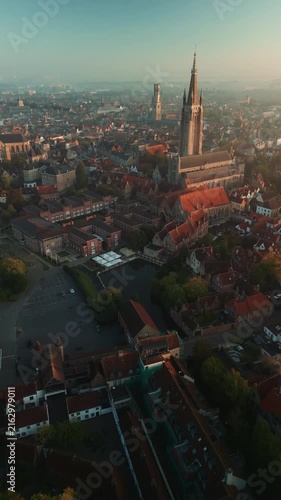Vertical drone panorama reveals Bruges, Belgium, with the Church of Our Lady, Belfry tower, red rooftops and historic center in morning haze. Vertical video.