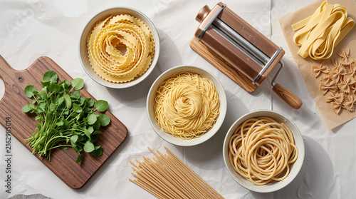 Flat Lay of Fresh Homemade Artisan Pasta Varieties with Vintage Pasta Maker Machine, Raw Ingredients, and Fresh Herbs on White Table