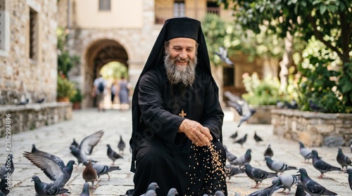 Smiling Orthodox Christian monk feeding pigeons in a monastery courtyard. Senior priest in black cassock with wooden cross scattering grain for birds