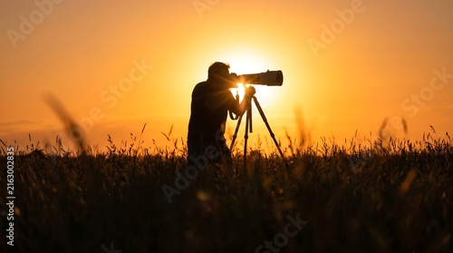 Silhouette of a photographer capturing a vibrant sunset over a grassy field with a tripod