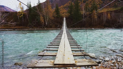 Wooden suspension bridge crosses a turquoise river. Mountains with autumn trees rise in the background. The bridge stretches forward, inviting travelers to cross