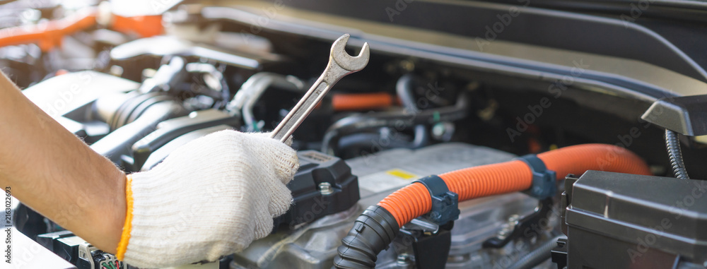 Fototapeta premium Auto mechanic hand holding a wrench during inspection and repair with electric hybrid car engine bay, professional vehicle maintenance service.