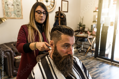 Konstfotografi Female millennial barber uses electric clippers to refine a bearded client's fade and sideburns in a modern salon