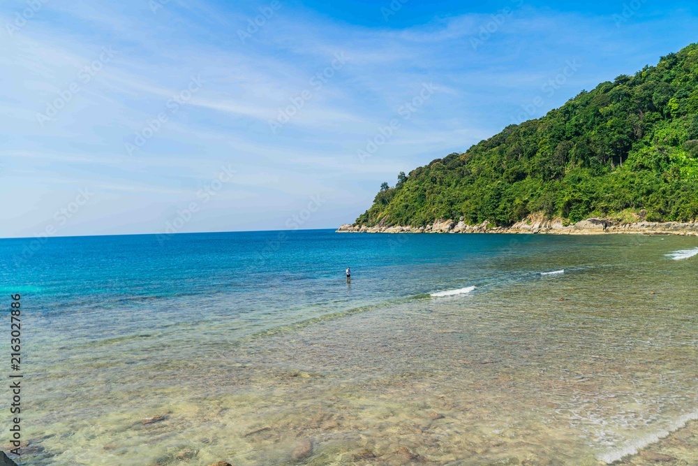Fototapeta premium The backdrop of the sky is a pristine tropical coastline with rocks, palm trees, turquoise sea, and a relaxing beach atmosphere. Merlin Snorkelling Beach, uncrowded Patong, Phuket, Thailand.