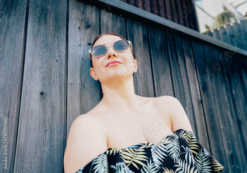Фотография Woman with short brown hair wearing sunglasses leans against a wooden wall, dres