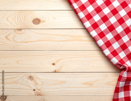 Red and white checkered tablecloth corner on light pine wood planks background, rustic picnic texture with copy space for food or text overlay design.
