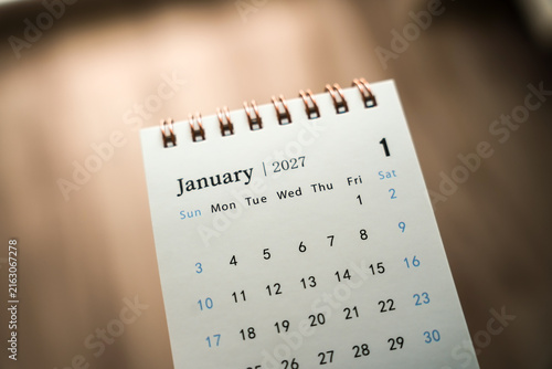 Small spiral desk calendar showing January 2027 dates on a wooden table with dramatic side lighting