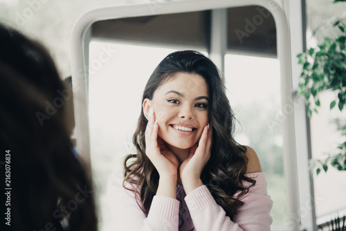 Cuadro en lienzo Young woman smiling at mirror while touching her face, confident self care and n