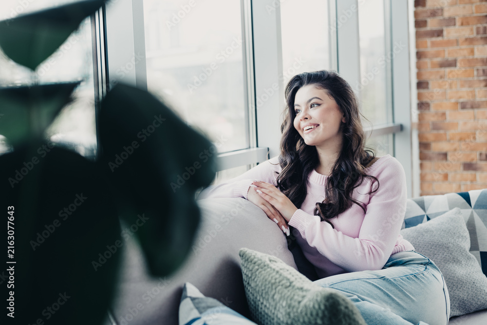 Obraz premium Young woman smiling and looking out window while relaxing on a cozy sofa in a bright living room