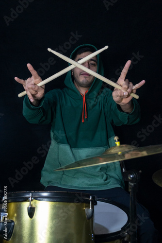 Obraz na płótnie Young drummer wearing a green hoodie posing with crossed drumsticks and making the rock and roll horns hand gesture