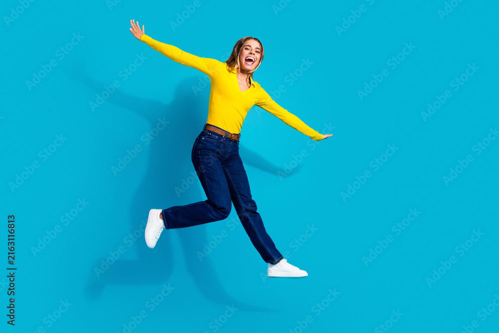 Fototapeta premium Joyful woman jumping against bright blue background wearing yellow sweater and jeans, energetic happy young adult celebrating with arms outstretched