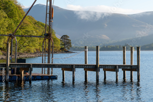 Wooden jetty on Derwentwater with calm blue water, sunlit woodland and mist drifting across Lake District mountains in Cumbria, England.