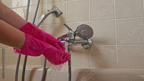 A hand in a bright pink rubber glove adjusts a chrome shower mixer in a beige-tiled bathroom; water streams from the fixture, illustrating the active cleaning or testing phase