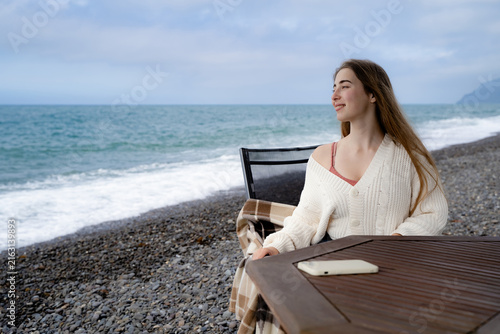 Canvastavla A young woman is relaxing alone at a seaside table, looking into the distance on a cloudy day