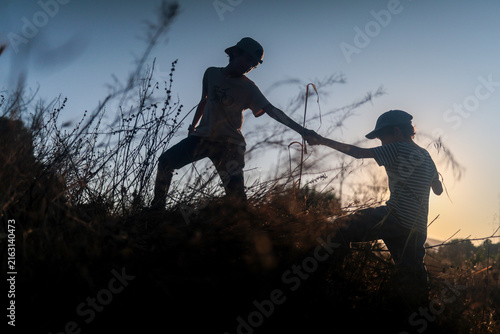 Quadro su tela Young boys helping each other climb a grassy hill at sunset, symbolizing teamwor