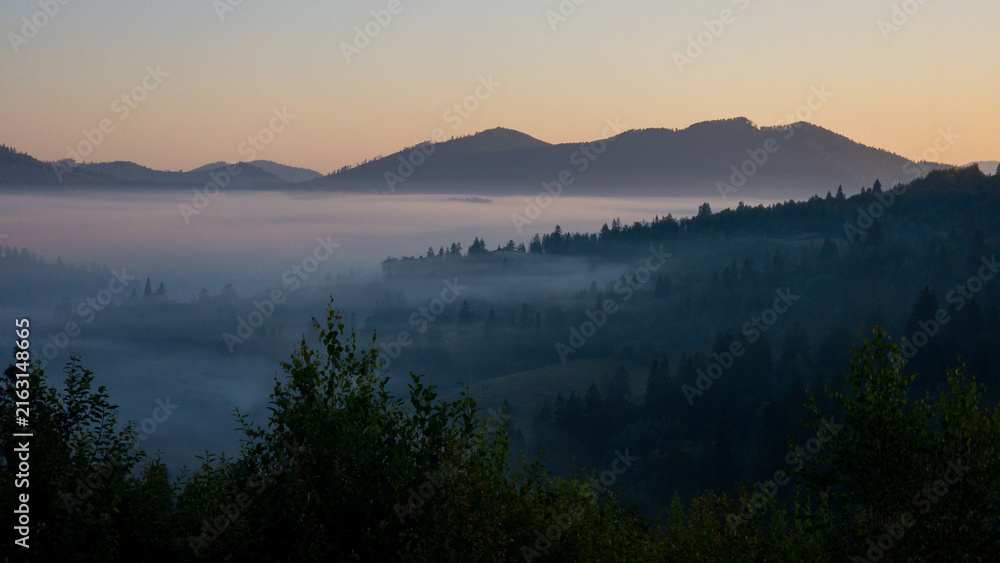 Fototapeta premium Scenic outdoor stock photo of a tranquil morning landscape featuring dense fog rolling over forested hills and distant mountain ridges under a soft dawn sky. Concept for serene nature, eco travel, and